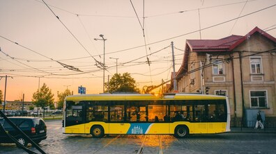 Жовтий низькопідлоговий тролейбус «Електрон» їде брукованою дорогою через трамвайні колії на тлі будинку з червоним дахом. Промені вечірнього сонця, що заходить, яскраво підсвічують вікна тролейбуса, а над дорогою розгалужується мережа контактних дротів