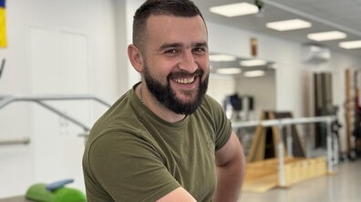 Захисник Богдан дивиться прямо в камеру та сміється. Він одягний у військову футболку зеленого кольору. Позаду видно різне реабілітаційне обладнання.