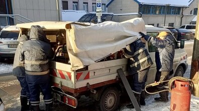 Зимова міська вулиця. Біля узбіччя припаркований невеликий службовий вантажний автомобіль із відкритим кузовом, накритим світлим брезентом. Навколо машини працюють четверо чоловіків у темному зимовому спецодязі зі світловідбивними смугами. У кузові видно технічне устаткування та інструменти. Праворуч на землі стоїть червоний газовий балон, від якого тягнеться шланг до обладнання в машині.