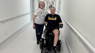 Пані Віра зі своїм сином Володимиром у просторому холі центру UNBROKEN: Володимир сидить у кріслі колісному, а пані Віра стоїть поруч. Обоє дивляться в камеру й усміхаються.