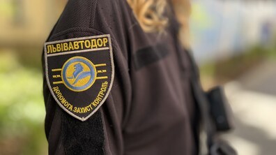 Великим планом зображено плече працівниці у темній формі з фірмовим шевроном «Львівавтодор», на якому вишито напис «Допомога, захист, контроль» та круглий жовто-синій логотип.