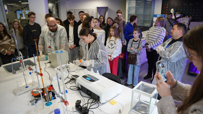 Заняття в лабораторії Lviv Open Lab. Навколо робочого столу з лабораторним обладнанням, колбами та вимірювальними приладами зібралася група дітей, підлітків і дорослих, які спостерігають за проведенням експерименту. У центрі кадру учасники в захисних халатах демонструють дослід, а частина відвідувачів фотографує процес на телефон.