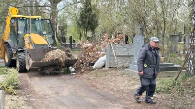 На фото кладовище, де тривають роботи з прибирання території. Жовтий екскаватор із ковшем вивозить купу сухого листя та гілок з алеї між могилами. Поруч проходить чоловік у робочому одязі, а навколо — надгробки, дерева та весняна зелень.