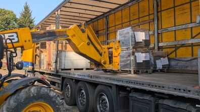 Жовтий екскаватор забирає з вантажівки спеціальне велике обладнання запаковане в ящики та поліетиленову плівку і картон.