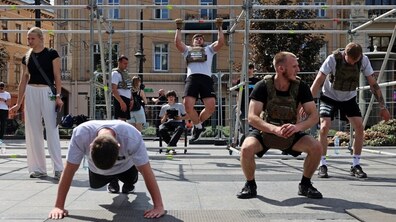 На площі у центрі Львова учасники спортивного заходу виконують фізичні вправи на спеціально облаштованому майданчику. Одні відтискаються, інші виконують підтягування та присідання. Позаду видно історичні будівлі міста та перехожих.