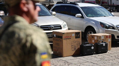 Військовослужбовець у формі стоїть на передньому плані. Позаду нього припарковані автомобілі, а на тротуарі лежать картонні коробки та кілька чорних валіз, у яких є обладнання, яке місто передає для військових. Знімок зроблено на одній з міських вулиць.