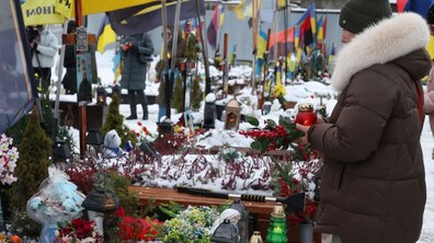 Жінка стоїть перед могилами полеглих Захисників на Личаківському кладовищі; в руках тримає лампадку та декоровану гілочку ялинки