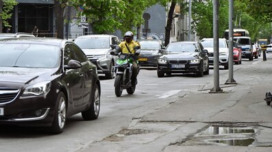 Вулиця Героїв УПА. У центрі їде мотоцикліст у жовтій куртці, довкола — щільний потік автомобілів. Асфальт потрісканий, біля тротуару видно калюжі. Обабіч дороги ростуть зелені дерева, праворуч — стовпи та світлофор.
