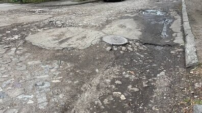 Частина вул. Юхновського з сильно зношеним дорожнім покриттям. Асфальт і бруківка місцями пошкоджені, на проїжджій частині є численні вибоїни, тріщини та нерівності. Посеред дороги виступає люк, поруч видно калюжі та розбиті краї покриття.