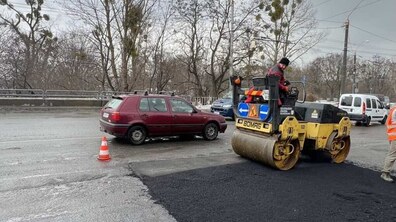 Ремонт дорожнього покриття на широкій ділянці дороги. На передньому плані — свіжо покладений темний асфальт, який ще виглядає вологим і рівним на фоні старого світлішого покриття. Праворуч працює дорожній каток — на ньому сидить чоловік у темному одязі та шапці, він керує технікою і ущільнює новий асфальт. На задньому плані — інші автомобілі, огорожа та дерева без листя.