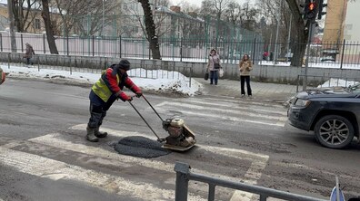 дорожній працівник ремонтує аварійну яму на проїжджій частині взимку. Узбіччя вкрите снігом і брудом, повз місце ремонту проїжджає автомобіль. Жінки стоять на узбіччі і чекають на зеленеий сигнал світлофора..