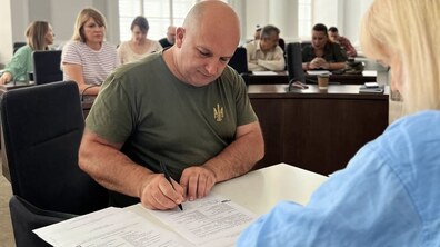 Ветеран у футболці кольору хакі із тризубом підписує угоду про грант від міста. На столі лежать документи. Позаду інші переможці гранту розділяють важливу мить одне з одним