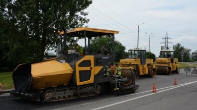 На дорозі стоїть спецтехніка: жовто-чорний асфальтоукладальник Volvo та дві великі дорожні машини для ущільнення покриття. Також видно дерева, лінії електропередач та робітників.