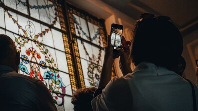 Люди розглядають, а одна жінка фотографує на смартфон великий сецесійний вітраж. Крізь матове скло пробивається м'яке світло, підсвічуючи витончені рослинні орнаменти та яскраві кольорові елементи візерунка.