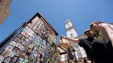 Один із торгових стендів Ветеранського ярмарку у Львові, де відвідувачі знайомляться з авторською продукцією. У кадрі покупці уважно розглядають асортимент нашивок, шевронів і сувенірів, спілкуючись між собою. Позаду видніється будівля міської Ратуші.