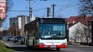 Великий низькопідлоговий автобус білого кольору з червоним бампером, який курсує за маршрутом №51 у Львові. Транспорт рухається міською вулицею, на другому плані - житлові будинки та автомобілі