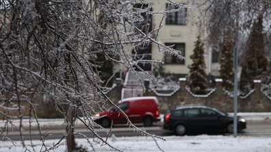 На передньому плані - вкриті прозорим льодом гілки дерев, крізь які на розмитому тлі видно засніжену вулицю з будинком та автомобілі