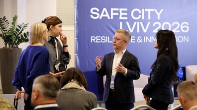 У залі на тлі синього банера з написом «SAFE CITY LVIV 2026» стоячи спілкується група учасників заходу. Чоловік в окулярах активно жестикулює, розповідаючи щось колегам.