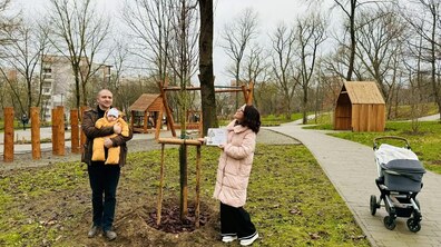 У центрі кадру — молоде тонке дерево, стовбур якого підтримується дерев'яною опорою. Навколо коренів свіжорозкопана земля, присипана мульчею (корою). Зліва від дерева стоїть чоловік. Він усміхається і ніжно тримає на руках немовля. Справа від дерева стоїть жінка в довгій світло-рожевій куртці та окулярах. Вона з посмішкою дивиться вгору на гілки дерева.