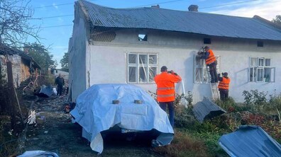 Комунальники у помаранчевих сигнальних жилетах закривають захисною плівкою пошкоджені вікна приватного будинку. На подвір’ї видно уламки, будівельне сміття та зняті частини покрівлі. Поруч стоїть автомобіль, накритий захисним тентом.