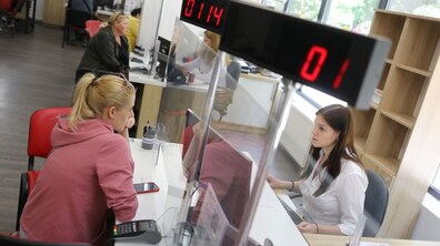 Працівниця Центру надання адміністративних послуг, сидячи на робочим столом, спілкується з відвідувачкою, яка сидить навпроти неї; позаду – ще одна працівниця спілкується з відвідувачкою; вгорі – електронне табло, яке показує черговість прийому