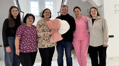 Спільне фото: у холі лікарні святої Анни стоять пацієнтка та її чоловік: чоловік тримає на руках немовля, загорнуте в білий коцик, а жінка тримає букет квітів. Поруч стоять дві медичні працівниці та двоє доньок подружжя. Усі щасливі та усміхнені.