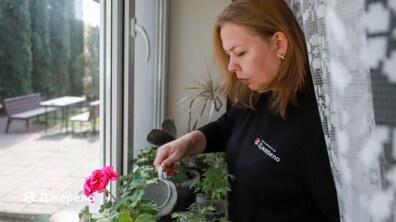 Соціальна робітниця «Джерела» Тетяна Олійник стоїть біля вікна і поливає кімнатні рослини з невеликої лійки. Жінка у квартирі старшої людини, за якою доглядає.