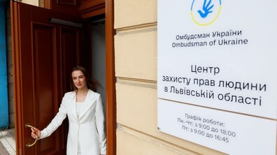 Молода усміхнена жінка відчиняє масивні дерев'яні двері будівлі, ніби запрошуючи увійти. Праворуч на стіні висить велика біла вивіска з офіційним синьо-жовтим логотипом та текстом: «Омбудсман України. Центр захисту прав людини в Львівській області», а також вказано графік роботи установи.