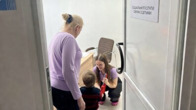 Соціальна працівниця з маленьким хлопчиком у кабінеті соціальних послуг. Вона привітно усміхається до дитини та тримає в руках іграшку. Поруч стоїть ще одна соціальна фахівчиня. Фото ілюстративне.