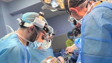 Троє лікарів оперують пацієнта, їм асистують медичні сестри; лікарі одягнені у спеціальні медичні халати, маски та окуляри