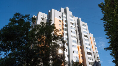 Висотна сучасна львівська багатоповерхівка білого та світло-помаранчевого кольорів. Будинок оточений зеленими деревами, а над ним – чисте блакитне небо. Яскраве сонячне світло пробивається крізь дерева та освітлює фасад будівлі. Фото – ілюстративне.
