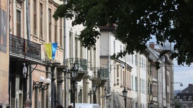 Багатоповерхові будівлі у центрі міста. На фасаді будинку видно український прапор, а поруч росте велике дерево, крона якого частково затуляють споруду. Вздовж вулиці є ліхтарі.