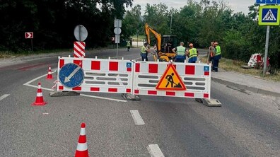 Ділянка дороги, перекрита бар'єрами з попереджувальними знаками. За огородженням видно групу працівників у жилетах та жовтий екскаватор на тлі дерев та похмурого неба.