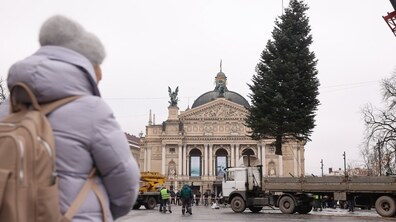 Момент встановлення новорічної ялинки перед Оперним театром у Львові. Посеред площі стоїть вантажівка з піднятою на тросі великою ялинкою. Декілька працівників у жилетах працюють навколо. На передньому плані — людина в світлій зимовій куртці з рюкзаком, яка спостерігає за процесом. На тлі — велична будівля Оперного театру з колонами та скульптурами.