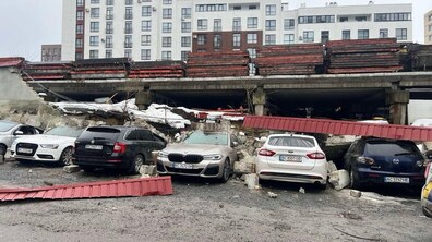 Відкрита автостоянка біля багатоповерхового житлового будинку. На передньому плані припарковані п’ять легкових автомобілів різних кольорів. Безпосередньо за автомобілями частина підпірної стінки обвалилася. Уламки конструкції та металеві елементи лежать на машинах і поруч з ними.
