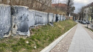 Пішохідна зона вулиці біля Органного залу у Львові. Уздовж схилу тягнеться стара бетонна підпірна стінка: поверхня стінки потріскана, місцями відшарована. Нижні частини окремих секцій сильно пошкоджені, видно оголений бетон і нерівності. Далі видно дорогу з припаркованими автомобілями, дорожні знаки та кілька будинків.