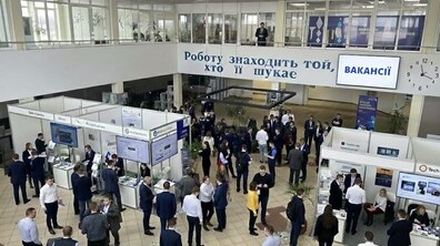 Великий хол центру зайнятості, де відбувається ярмарок вакансій. Люди спілкуються біля стендів компаній, знайомляться з пропозиціями роботи та обговорюють можливості працевлаштування. Більшість учасників одягнені в діловому стилі. На стіні видно написи: «Роботу знаходить той, хто її шукає» та «ВАКАНСІЇ»
