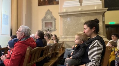 Слухачі різного віку сидять на дерев'яних лавах у величній залі з класичним інтер'єром та уважно дивляться в бік сцени. На передньому плані праворуч сидить жінка, яка тримає на руках маленьку світловолосу дитину, а ліворуч видно чоловіка в яскраво-червоній куртці та інших відвідувачів.