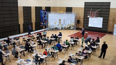 Великий спортивний зал облаштований під шаховий турнір, де десятки дітей сидять за окремими столиками, зосереджено граючи партії. На дальній стіні розміщено великий банер із написом «Шаховий Львів» та зображенням шахових фігур