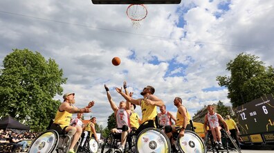 Динамічний момент матчу з баскетболу на кріслах колісних, який проходить на відкритому майданчику під хмарним небом. Гравці у жовтій та біло-червоній формі із написом «RIVNE» борються під кільцем за м'яч, що летить у повітрі.