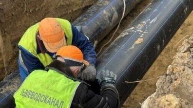 Двоє працівників у помаранчевих касках та світловідбивних жилетах із написом «Львівводоканал» працюють у глибокому земляному котловані. Вони схилилися над великою чорною пластиковою трубою, виконуючи монтажні роботи. Поруч у траншеї прокладено ще одну таку ж трубу.