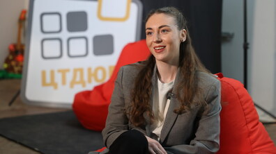 Роксолана Шиманська сидить у студії на червоному кріслі-мішку та усміхається під час розмови. На ній сірий піджак і світла футболка, а до одягу прикріплений мікрофон. Позаду видно елемент декорації з написом і розмите студійне тло.