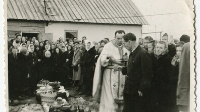 Великдень на засланні, Інта, Комі АРСР (тепер Республіка Комі, Російська Федерація), 1957 р. У центрі фото стоїть священик у світлому святковому облаченні, розшитому хрестами. Він тримає кропило, освячуючи дари. Перед ним стоять люди, зокрема чоловік у темному пальті, схиливши голову. На землі довгою вервицею виставлені плетені кошики та вузлики з продуктами, накриті вишитими рушниками. На задньому плані видно великий натовп людей. Вони стоять біля простої сільської будівлі з дерев’яним дахом, на якому закріплена драбина. Фото з архіву Ярослави Гасюк.