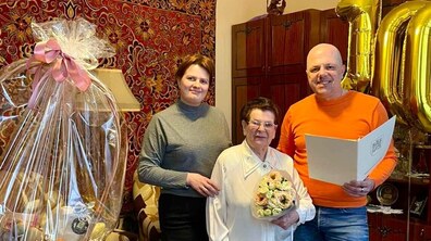 Ганна Сопрунова (по центру) з букетом квітів, позаду видно золоті повітряні кульки з цифрою “100”. Ліворуч на столі стоїть великий подарунковий кошик, а інтер’єр кімнати з килимом на стіні та дерев’яними меблями створює затишну, родинну атмосферу свята.