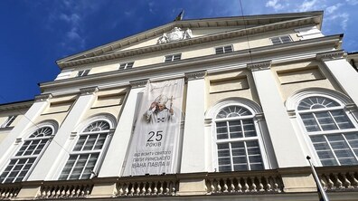 На фасаді будівлі Львівської міської ради висить великий банер із зображенням Папи Римського та написом: «25 років від візиту святого Папи Римського Івана Павла ІІ». Над будівлею видно яскраве синє небо з білими хмаринками.