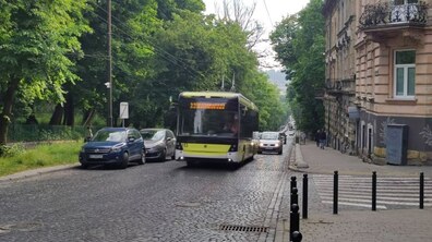 Тролейбус №22 їде однією з вулиць міста. Поруч їдуть автівки, вздовж дороги видно зелені дерева та фасад будинку.