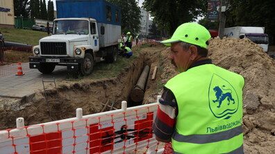Ремонтні роботи по заміні водопроводу на вул. Зелена. Вздовж дороги викопано глибоку траншею з великою трубою всередині. Праворуч стоїть працівник у яскравому жилеті «Львівводоканалу» та спостерігає за роботами. На задньому плані видно дерева, будівлі та автомобілі.