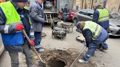 міська вулиця, де посеред дороги є великий круглий отвір у асфальті — відкритий каналізаційний колодязь без кришки. Навколо нього працюють п’ятеро працівників Львівводоканалу