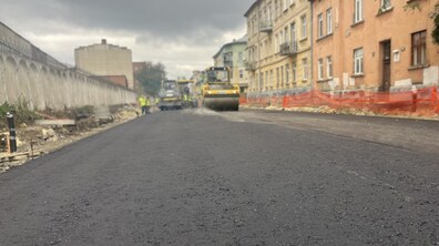 На вул. Залізничній триває асфальтування проїзної частини. На свіжоукладеному темному асфальті працює дорожня техніка, зокрема жовтий дорожній каток. Обабіч дороги облаштовують тротуари, ділянки робіт огороджені помаранчевою сіткою.