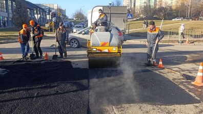 Ремонт дорожнього покриття методом укладання гарячого асфальту. У центрі кадру – спецтехніка жовтого кольору, яка ущільнює щойно покладений чорний асфальт. Від свіжого покриття піднімається пара, що свідчить про високу температуру суміші. Ліворуч стоять троє працівників у робочому одязі сірого кольору з яскравими помаранчевими вставками та у помаранчевих касках. Праворуч ще один працівник з лопатою контролює край ділянки.